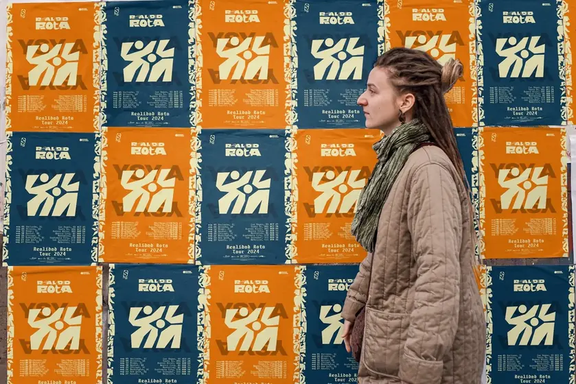 Wall covered in orange and blue Realidad Rota Tour 2024 posters, with a woman in profile in front