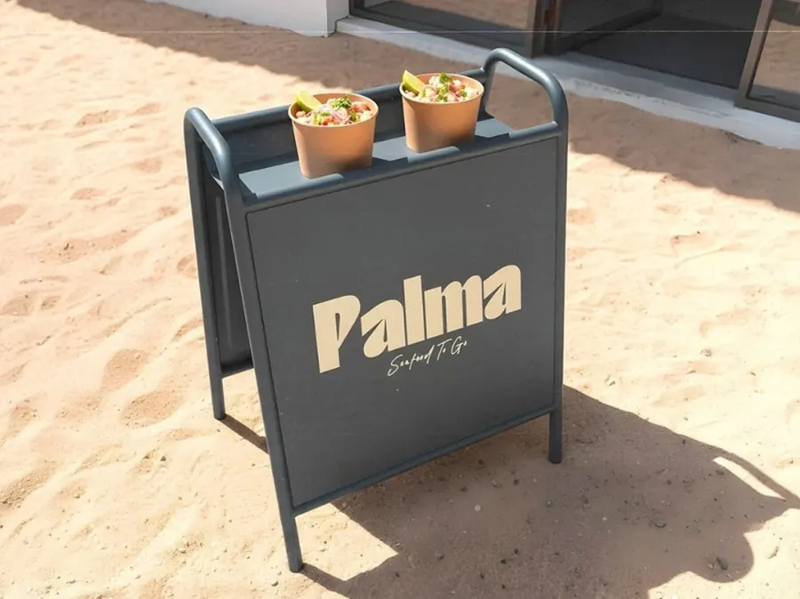 Sidewalk sign for Palma Seafood To Go standing on sand, with two cups of food on top, by CBAS